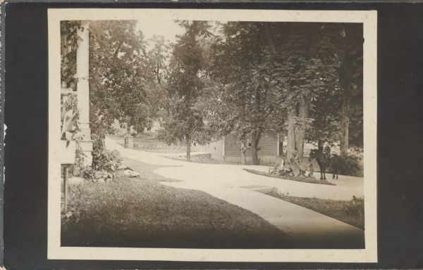 Postcard of the new cement driveway. The corner of the house is in the left foreground, and at the entrance to the drive is a gate and fence. On the right a young child is sitting on horseback on the turn of the driveway near a tree. Behind the tree is a building, perhaps a garage. Text on back of postcard reads: "I am sending you several cards of the new cement driveway to the house. Thinking you would like to see what it is like. This view shows the turn in front of the house. We are very proud of this improvement. With love from us all to you. Irene Searson, July 14, 1909." Addressed to Mrs. McCormick, 135 Rush St., Chicago, Ill.