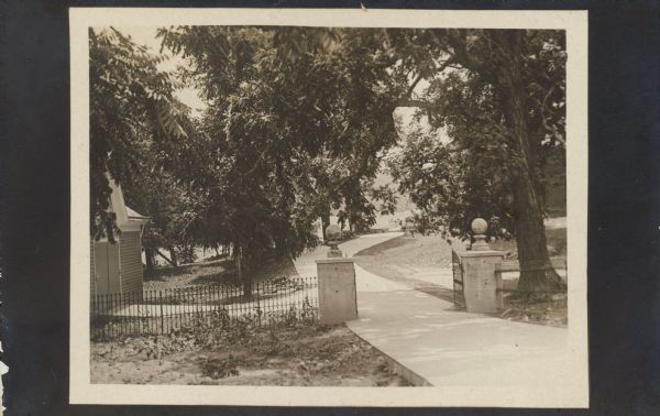 View looking towards the gate and fenced entrance of the new cement driveway. A building, perhaps a garage, is behind the fence on the left. The house is behind trees on the right. Text on back of postcard reads: "This card shows the new cement drive way to the house, looking from the road. It is a great improvement to Walnut Grove, and Mr. Searson spent many hours planning as to how it would be best made etc. July 14, 1909." Addressed to Mrs. McCormick, 135 Rush St., Chicago, Ill.