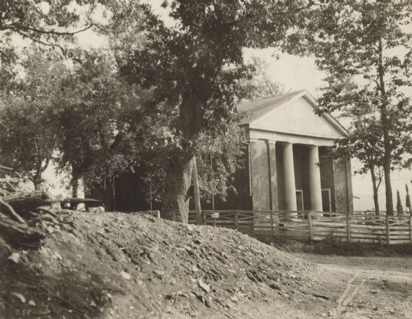 View toward the entrance to the manor house at Walnut Grove. A fence surrounds the house, and a man is standing near an automobile parked on the left behind a small mound of dirt or a hill.