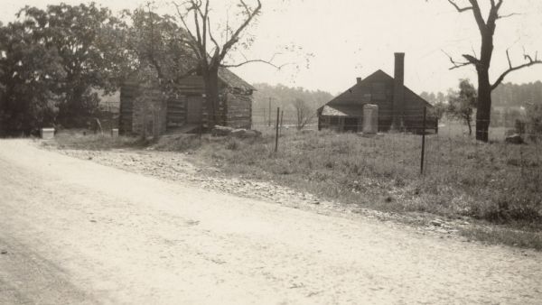 View from fence-lined road towards two buildings. The log building on the left has an exterior chimney to the left of the door, and the door has a wooden walkway to it from the road. Behind the fence in the center is what may be a stone marker on a slab, and behind it is another building with an exterior chimney  on the right, and a small porch on the left. Text on back reads: "The old workshop on the McCormick Farm in Virginia (near Raphine) where Cyrus McCormick worked on his reaper."