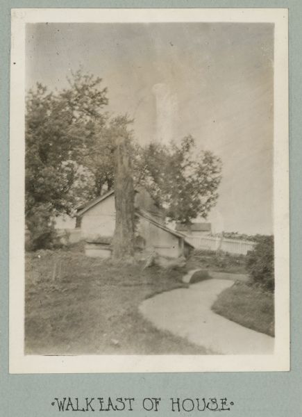 Photograph from album with the caption: "Walk East of House" which shows a sidewalk curving towards a building. A fence and other buildings are among trees in the background.