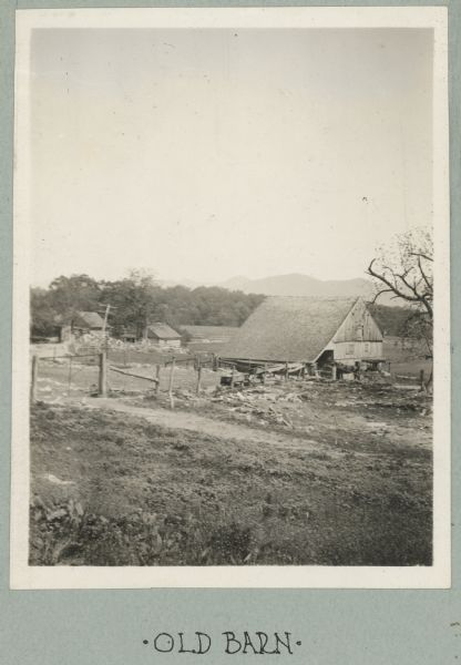 Photograph from album with the caption: "Old Barn." The view is down a hill towards the barn which has a fenced enclosure. A horse-drawn buggy is near the barn on the right. In the background on the left are two small log buildings. Mountains are in the distance.