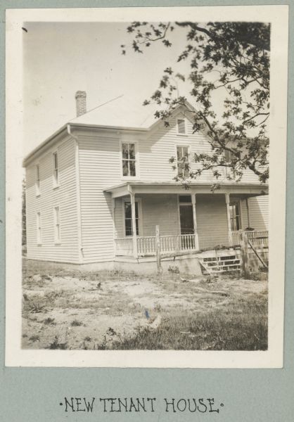 Photograph from album with the caption: "New Tenant House." View towards the front of the two-story house which has a porch. There is a wire fence with wooden posts in front.