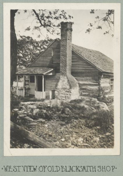 Photograph from album with the caption: "West View of Old Blacksmith Shop." View towards the log building, which has an exposed chimney on the right side, and a small porch on the left. There is a closed window above the porch near the peak of the roof. Large rocks are in the yard in front.