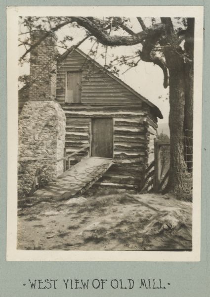 Photograph from album with the caption: "West View of Old Mill," with a view towards the front of the log building. There is a wooden walkway with a railing on the left that leads to the door on the right side, and an exposed chimney is on the left. On the second floor near the peak of the roof is a closed wooden door or window. The building is built into the side of a hill.