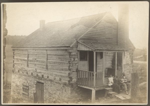 Three-quarter view towards the front and left side of a log building with a peaked roof which is built into a hill. The left side of the building is below the hill and the stone foundation is exposed. There is a door and window in the foundation. At the front is a porch on the left side, an exposed chimney on the right side, and a window with wood covering above the porch near the peak of the roof. A man, woman and young child are sitting together on the front steps. The man is holding a stringed instrument in his lap. Caption on back reads: "This is the shop, and I like it much better than those Mr. Murry(?) took. Alex & his daughter are on the porch. This shows a different front(?) from the one you have."