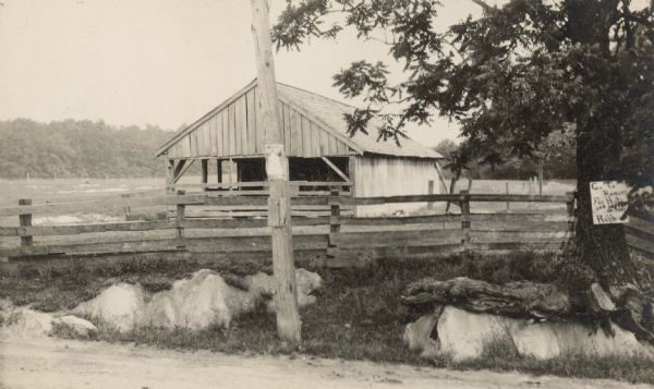 View from road towards a fence and a shed at Walnut Grove. Fields are in the background surrounded by trees. A sign on the tree in the right foreground reads, in part: "Got to Ronans(?) for Hats and Shoes Raphine." Handwritten on back: "Shed which Mr. Searson wants to move to build the cottage for darky. This is across the street and a little toward town from the Machine Shop."