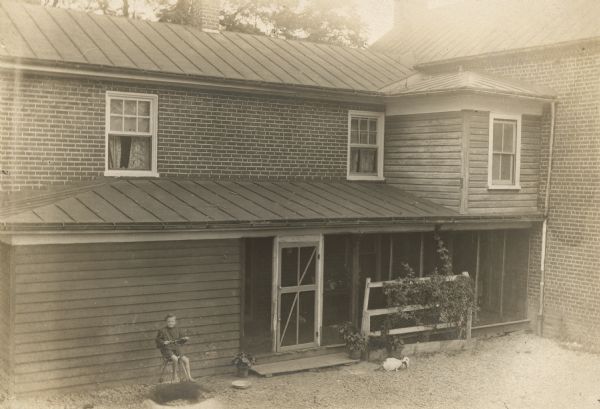Slightly elevated view looking at side of manor house with porch. A boy is sitting on a tricycle on the left, and two dogs, blurred by movement are moving in front of him. Cats are eating from a bowl in the yard near the porch door.