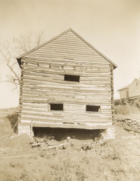 View looking up towards the back of the log mill building, which is built into the side of a hill. There are three small window openings. Two wheelbarrows are in front of the opening near the base of the stone foundation, and a man is working underneath the building. A building on the hill is in the background on the right.
