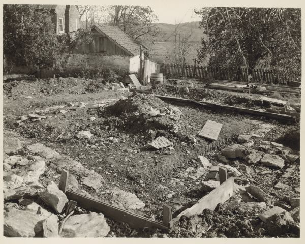 View of old foundation and debris. A fence, small building and the Walnut Grove brick manor house are in the background on the left.
