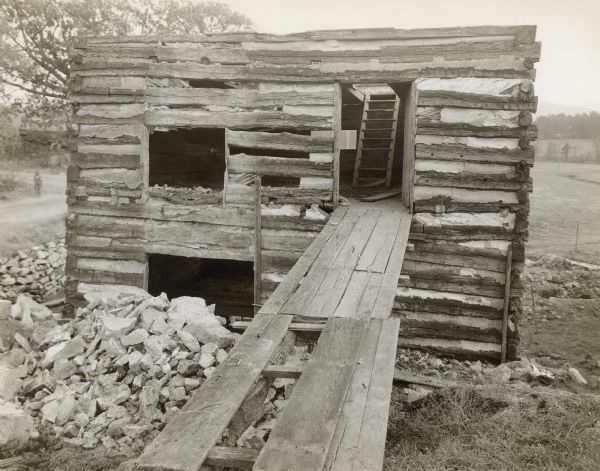 View down wooden walkway towards the front of the mill, which is built into a hill. The roof has been removed, and the interior stairway to the second floor can be seen through the open doorway. There are windows on the left on the first and second floors.