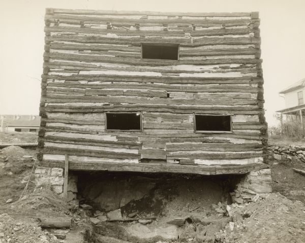 View looking up towards the back of the log mill building, which is built into the side of a hill. There are three small horizontal openings in the log wall, and a large opening near the base of the stone foundation. The roof has been removed. A building on the hill is in the background on the right, and another building, probably the barn, is in the background on the left.