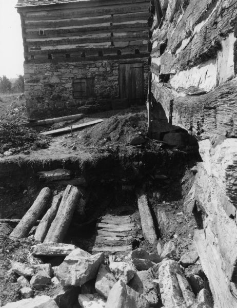 Handwritten on back: "Excavation in water wheel pit back of old mill building. Note three logs used as support for stone pillar to hold outer end of water wheel shaft. Also back boards of a portion of the old water wheel, which had rotted and fallen there and during the years, a hundred or more, had become covered with about five feet of dirt." There is another log building in background, probably the Blacksmith Shop.