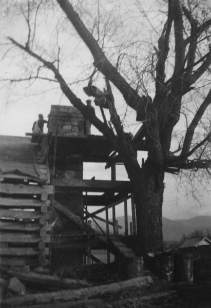 Men are standing on scaffolding working on the roof near the top of the chimney. A tree is near the log building on the right, and mountains are in the distance. Handwritten on back: "Slave Quarters in process of Restoration."