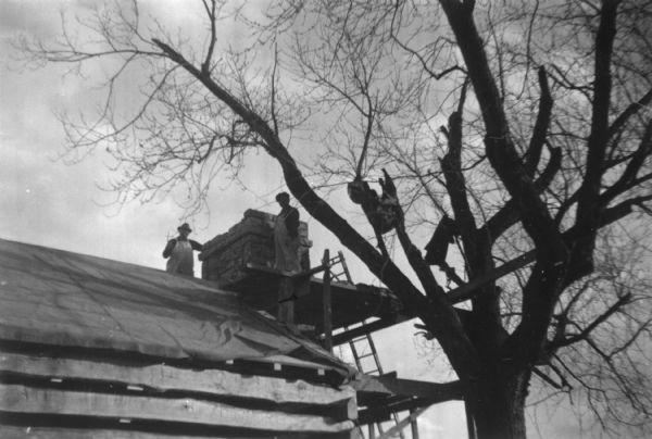 Men are standing on scaffolding working on the roof near the top of the chimney. A tree is near the log building on the right. Handwritten on back: "Slave Quarters in process of Restoration."