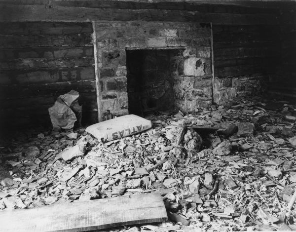 Interior view of stone fireplace downstairs. There are andirons inside the fireplace. Debris is on the floor. There is an unopened bag of Atlas cement near the left side of the fireplace.