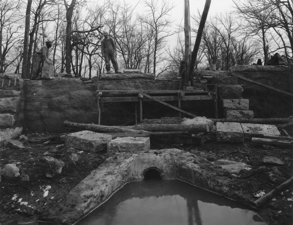 Two men are standing on the stone wall, and another man is standing on scaffolding inside the dam. In the foreground are large stones, logs, and water. Handwritten on back: "Detail of construction of inside of dam at breast wall or center.