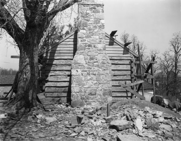 View of exterior chimney at end of slave cabin. Scaffolding is along the side of the building on the right, and stone and wood debris is in the foreground. There is a tree next to the corner of the building on the left. A fence and farm buildings are in the background. Handwritten on back: "Slave cabin. Note chimney at end of cabin and chinking between logs."