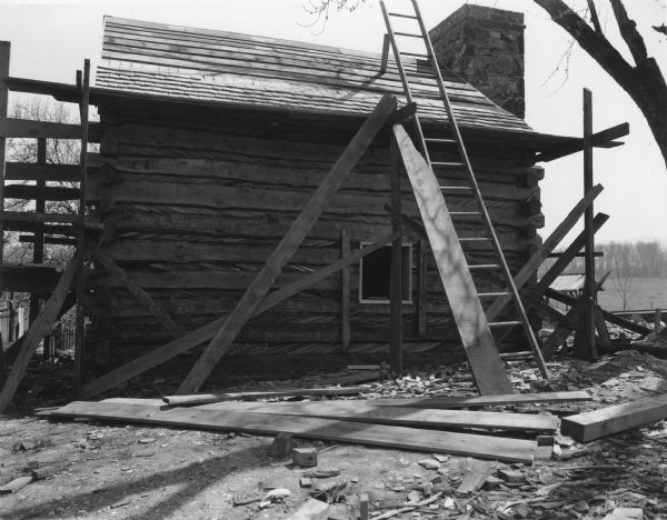 View towards side of slave log cabin which has a peaked roof. A ladder is set up for access to the roof. Scaffolding is on the left and right ends of the building. The exterior stone chimney is on the end of the building on the right. Handwritten on back: "West side of slave cabin — Note window frame."