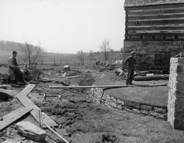 Two men are looking toward the back of the mill building which is on the right. Behind the man on the right is the Blacksmith Shop. In the distance are fenced-in fields and trees. Handwritten on back: "Tail race to carry off water which goes over wheel."