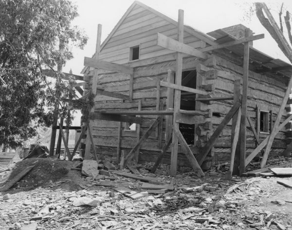 View towards front and right side of the log building with peaked roof and brick chimney. The front has a door and window opening, and another window in the peaked roof. Another window is on the right side of the building. The exterior stone chimney at the back of the building rises above the roof. Scaffolding is set up around the building. Handwritten on back: "North end of slave cabin. Note chinking between logs."