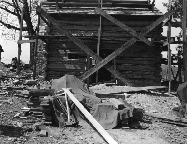 View towards side of cabin, which has a door and window opening in the log structure. Scaffolding is set up around the building, and tools and building materials are in the foreground. The exterior stone chimney is on the left side on the end of the building. Two men are sitting in the background on the left. Handwritten on back: East side of slave cabin. Note home made shingles on roof."