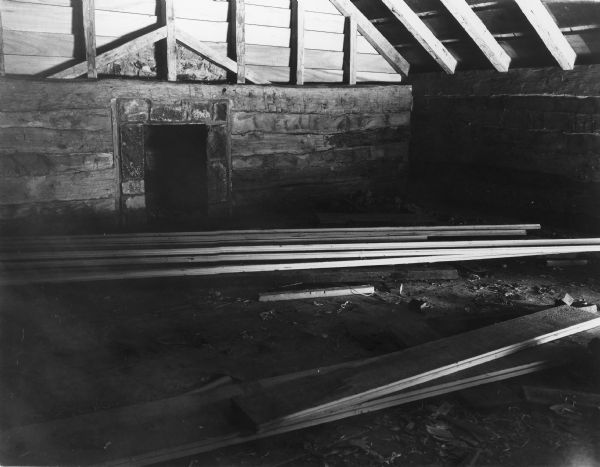 Interior view of log cabin upstairs. A small fireplace is on the far wall at the end of the cabin, and the peaked roof of beams and wood is exposed. Lumber and debris are on the floor. Handwritten on back: "Cabin upstairs."