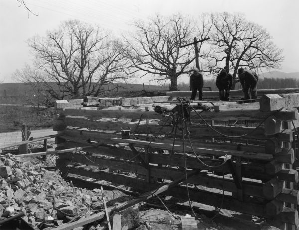 Three men are working on the top of the mill building. There is no roof, and the logs are not filled in or chinked. The road curves from the left in the background. Mountains are in the distance.