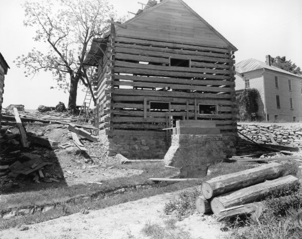 View looking up towards the back of the mill building with stone foundation built into the side of the hill. The mill is partially constructed of logs, not yet chinked, and has a peaked roof. A ladder is leaning on the back of the building, and scaffolding is near the roof. On the left is the corner of the Blacksmith Shop, and there is a building in the background on the right. Large logs are piled in the foreground. Handwritten on back: "Note tailrace, pillar to support outer end of shaft of water wheel, logs on which shaft rests, and opening in wall where shaft goes through."