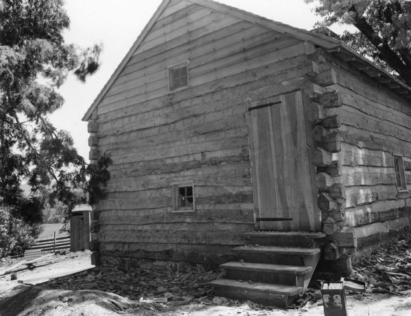 View towards the front and right side of the slave cabin built of chinked logs with a peaked roof. Steps lead up to the door, and a small window is on the left. There is a window above near the center of the peak. On the right side of the building is a small window. In the background on the left is a small outbuilding near a fence, and a field and mountains beyond. Handwritten on back: "End of slave cabin with door to second floor. Note strap hinges."