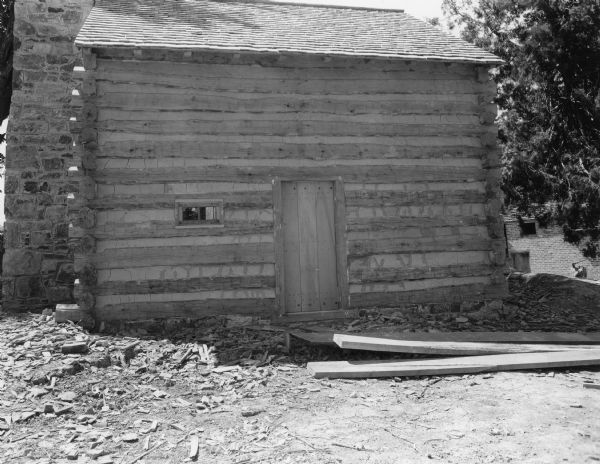 View towards side of slave log cabin with a door in the center and a small horizontal window to the left. The roof is peaked and shingles are in place. The exterior stone chimney is at the end of the building on the left. Debris and lumber are in the foreground. In the background on the right is a brick building, and in front of it is an axe on a block of wood. Handwritten on back: "Side of slave cabin showing door to first floor and side view of chimney."