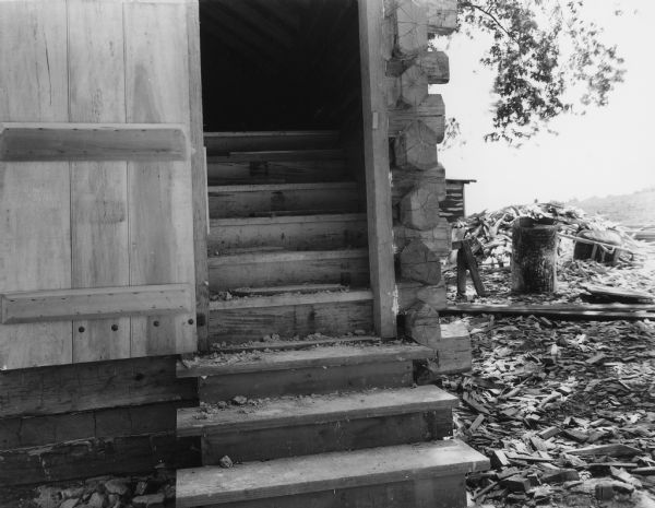 Exterior view of the open door at the end of the slave cabin. The steps continue to rise up inside the cabin to the second floor which has an exposed peaked roof. To the right of the cabin is a pile of wood, a wheelbarrow, and a small outbuilding. Handwritten on back: "Stairway to second floor of slave cabin. Note hand made nails in door."