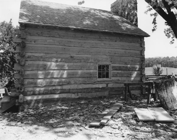 View of side of slave cabin with a small window. Steps are on the end of the building on the left. An exterior stone chimney is on the end of the cabin on the right and rises above the peaked roof which is shingled. In the foreground is debris, sawhorse and a barrel. A fence, hill and trees are in the background on the right.