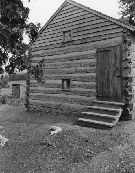 View towards the front of the slave cabin built of chinked logs with a peaked roof. Steps lead up to the closed door, and a small window is on the left. There is a window above near the center of the peak. In the background on the left is a small outbuilding near a fence, and a field beyond. Handwritten on back: "Exterior of Slave Cabin, north end, showing entrance to upper floor. Note hand wrought iron hinges and latch."
