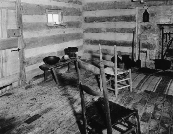 Interior view of a corner of the slave cabin which has a brick floor. There is a door on the left, a window to the right of the door, and a fireplace on the right. Handwritten on back: "Interior of the Slave Cabin, first floor, south east corner. Note wooden bench, iron basin, gourd dipper, wooden water bucket. Also door on east wall showing hand made lock covered with hand made wooden casing and hand made wrought iron latch. On small shelf under window home made lye soap on a forked stick."