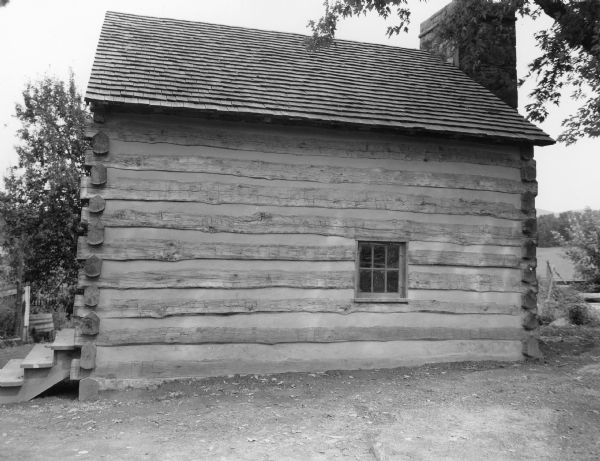 View towards side of slave log cabin which has a small window. Steps are on the end of the building on the left. An exterior stone chimney is on the end of the cabin on the right and rises above the peaked roof which is shingled. Handwritten on back: "Exterior of Slave Cabin, west side, note hand made shingles and wooden pegs to fasten window and frame. Also daubing between logs. This daubing is made of a mixture of lime, sand and white oak clay, according to an old formula."