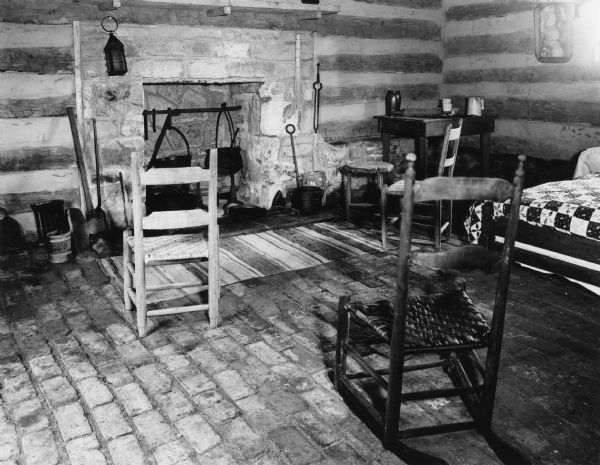 Interior view of a corner of the slave cabin which has a brick floor. A fireplace is on the far wall, and a bed is on the right. Handwritten on back: "Interior of cabin, first floor, southwest corner."