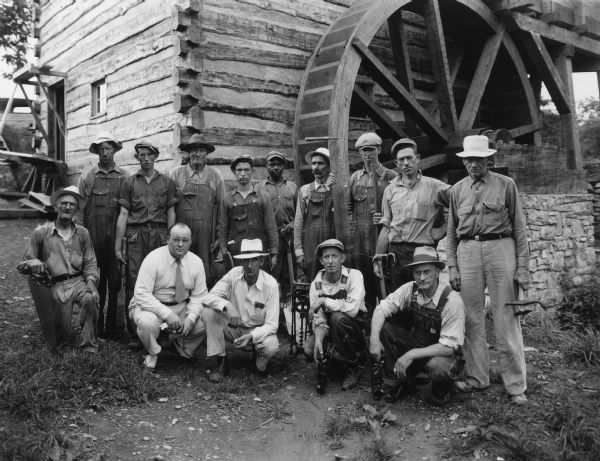 Group portrait of workers posing at the back of the Grist Mill at Walnut Grove. A number of the men are holding their tools. Herbert A. Kellar, in charge of the work, is crouching in the front row on the left wearing a shirt and tie. Handwritten on back: "Men at Walnut Grove Resoration, May to September 1938. Most of these men worked continuously in this period and some of them were on the job from the beginning to September 1937." A list of the men's names and where they are from is included.