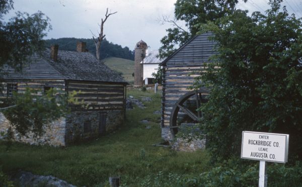 View towards the Grist Mill and Blacksmith Shop at Walnut Grove. In the background is a barn with a silo. A sign on the right reads: "Enter Rockbridge Co. Leave August Co."