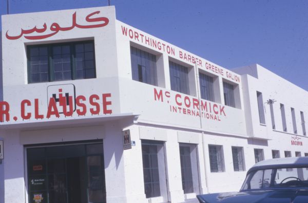 Exterior view of a white building with red signs that read, in part: "Barber Greene Galion," and "McCormick International." A sign above the entrance on the left reads, in part: "R.Clausse."ar
