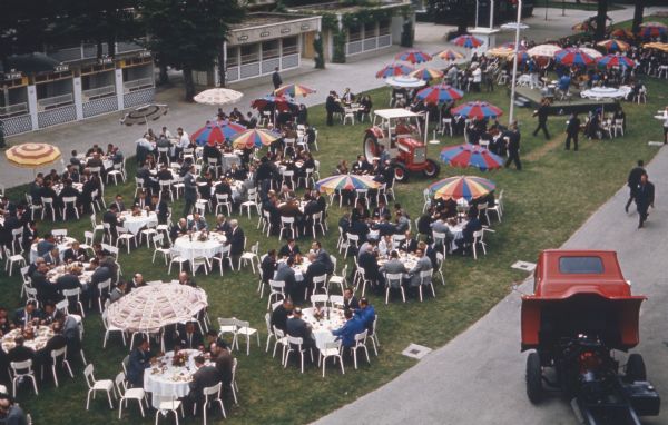 Elevated view of groups of men sitting and eating at round tables, some covered with umbrellas, on a lawn. A truck and a tractor are being exhibited on the grounds. In the upper right is a band on a low stage.