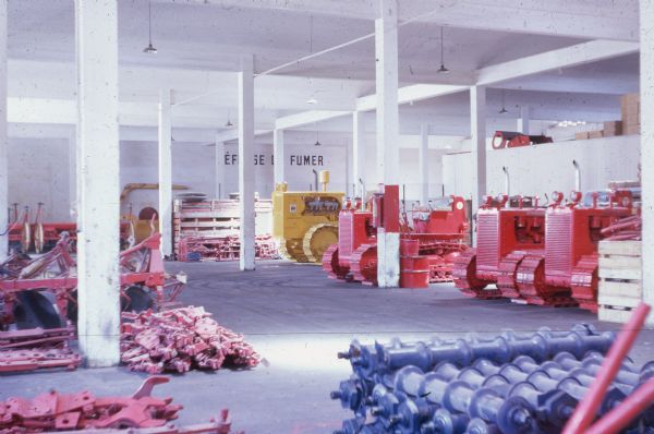 Agricultural equipment and parts inside a building. There are continuous track tractors parked in a row, one painted yellow, and three painted red. A sign on the back wall, blocked by pillars, reads: "Défense de Fumer."