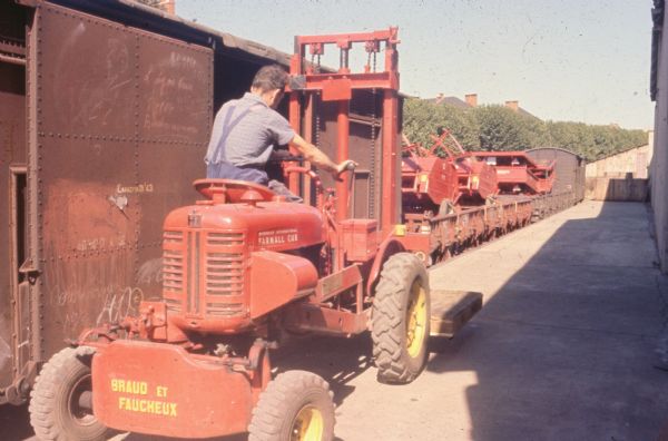 A man is working outdoors with a Farmall Cub with a forklift on a loading dock near railroad cars.
