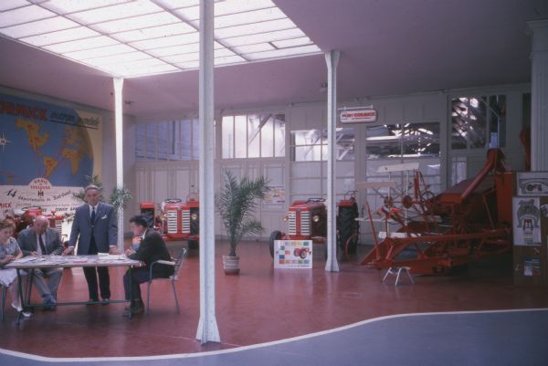 A group of people are working at a table on the left in a showroom. On display are tractors and a harvester. A large map of the world is on the wall in the background on the left, with text at the top that reads: "[McC]ormick marque mondiale." 