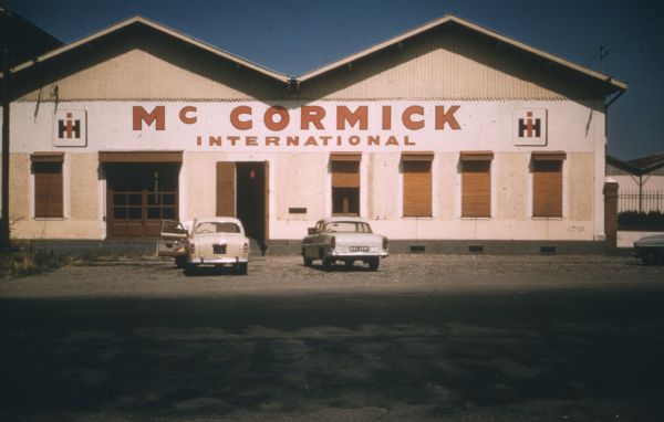 Two cars are parked in front of a building with a sign and a logo for International Harvester, McCormick International.