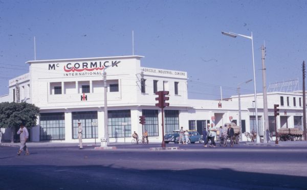 Exterior view across intersection towards a large white building with red signs that read: "McCormick International," and "Agricole, Industriel, Camions." Pedestrians, automobiles and horse-drawn wagons are on the street.