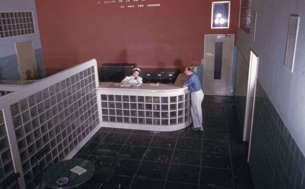 Elevated view of reception area in office. A woman wearing a white coat and hat is behind the desk on the telephone. A man is standing at the desk on the right. The sign on the far wall reads: "La Qualite de la vie est Faite du Prix des Hommes."