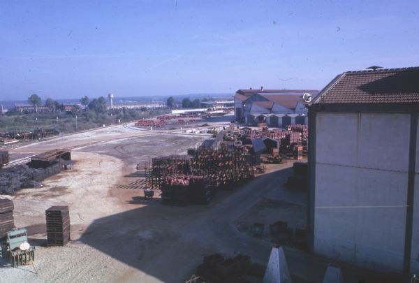 Elevated view of factory buildings, and tractors and other agricultural equipment parked on the grounds. A tower is in the distance, and tree-covered hills are in the background.