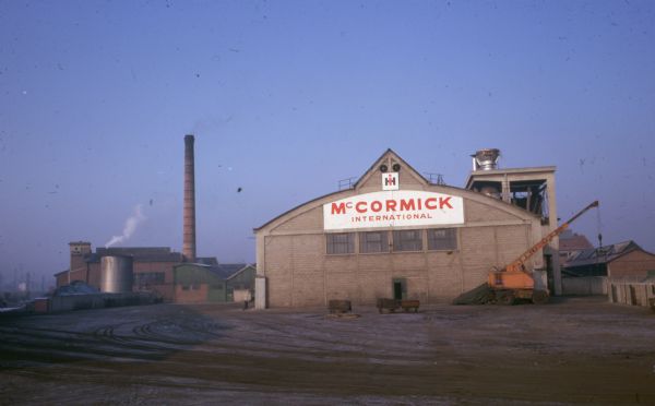 View toward a building with a large sign that reads: "McCormick International." The IH logo is above. A crane is at the corner of the building on the right, and in the background on the left are large factory buildings and a smokestack.