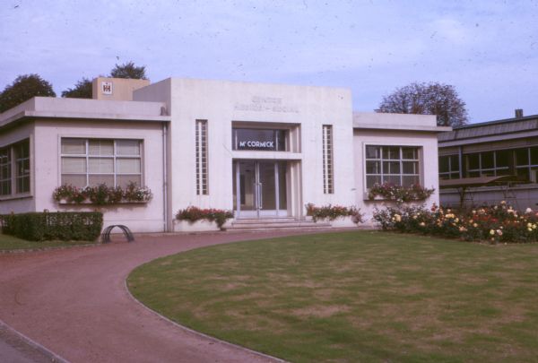 View toward the front of a building with a sign above the door that reads: "Centre Medico. — Social." A sign on the window above the entrance reads: "McCormick." The IH logo is on a wall in back of the building.